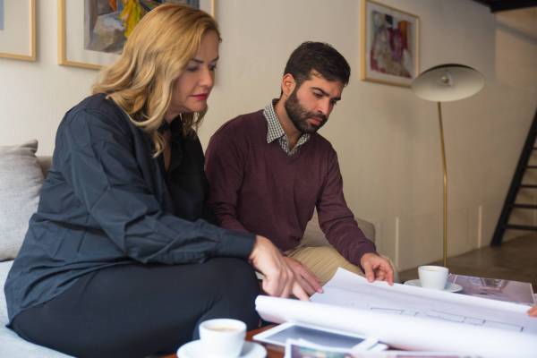 Interior designer and client studying blueprint and discussing renovation project. Young man and middle aged woman sitting on couch at home and talking. Renovation concept