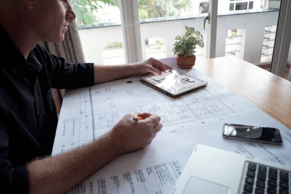 Side view of professional mature male architect drawing house blueprint and using digital tablet while sitting at office table